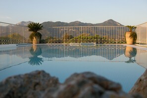 A clear swimming pool with a view of mountains in the background, surrounded by a white fence. - Residence Verdeluna (Pietrasanta)