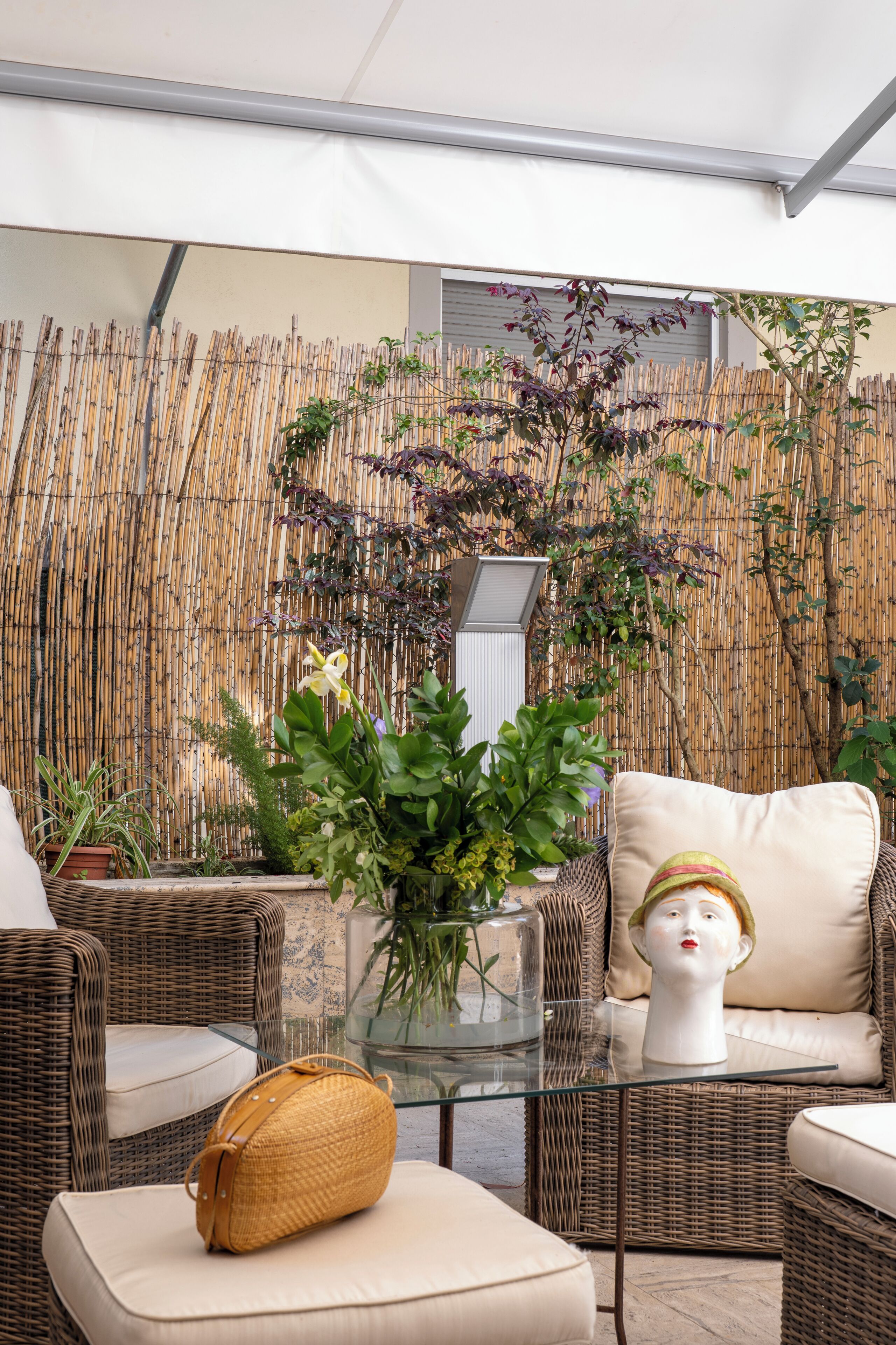 Un fauteuil de jardin en osier, une table en verre avec un panier tressé, et une plante en pot.