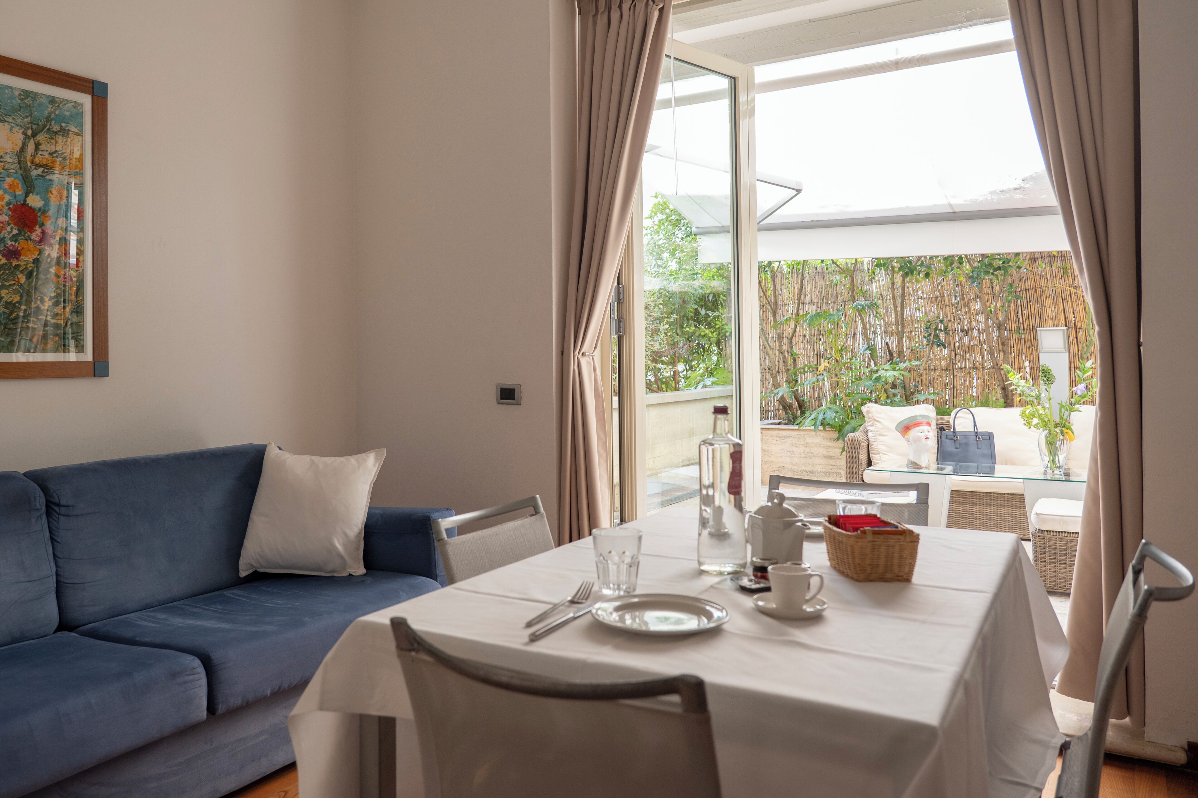 A dining area with a table set for two, a blue sofa, and a view of a garden through a glass door.