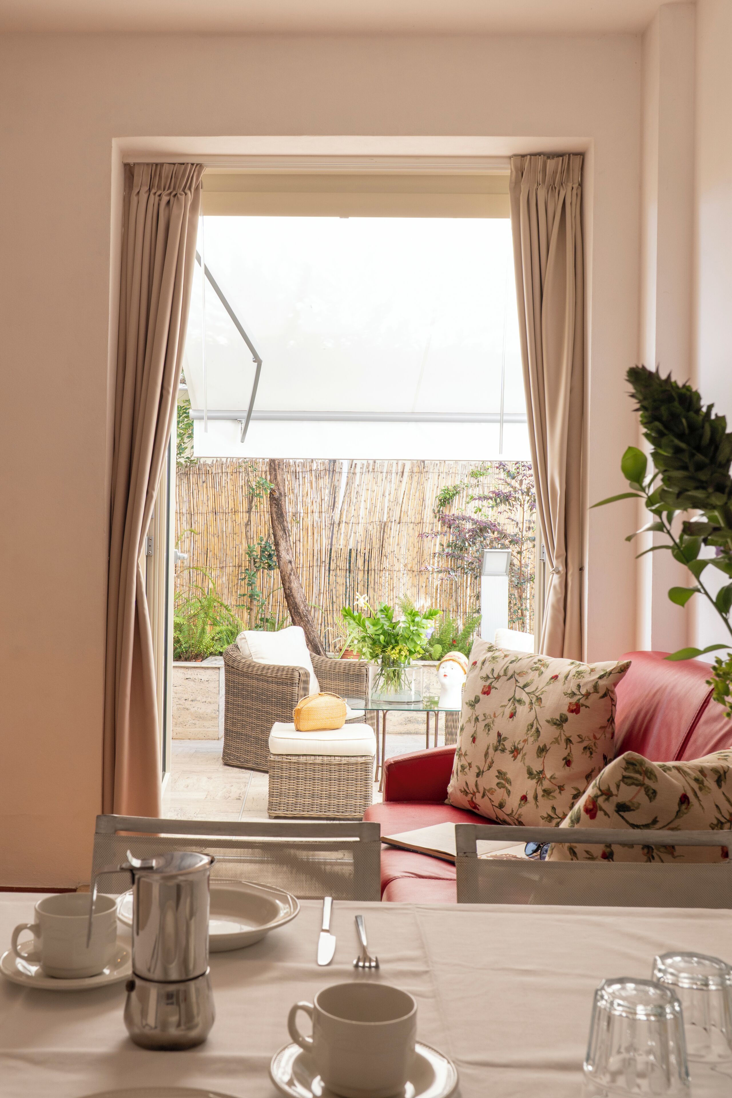 A room with a dining table set for two, a wicker sofa with floral cushions, and a view of a bamboo fence and plants outside.