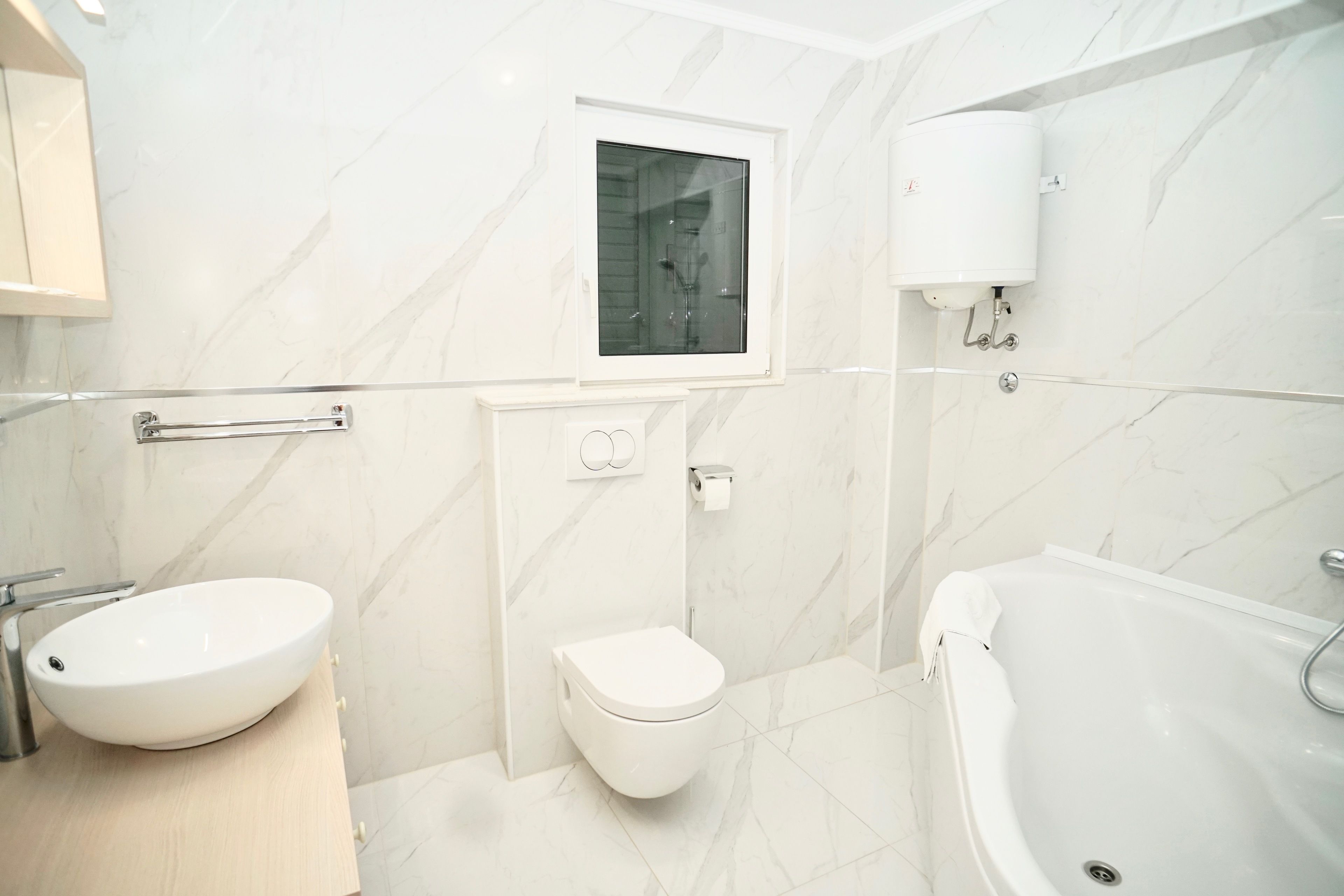Een moderne badkamer met een witte wastafel, toilet en badkuip, marmeren wanden en een raam boven het toilet.