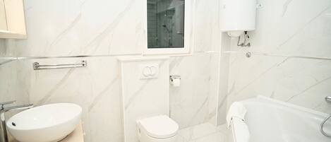 Een moderne badkamer met een witte wastafel, toilet en badkuip, marmeren wanden en een raam boven het toilet.