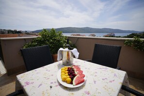 A table with a floral tablecloth, a pitcher, and a plate of fruit on a balcony overlooking a body of water and hills. - Villa Rustica Dalmatia Apartments (Seget)