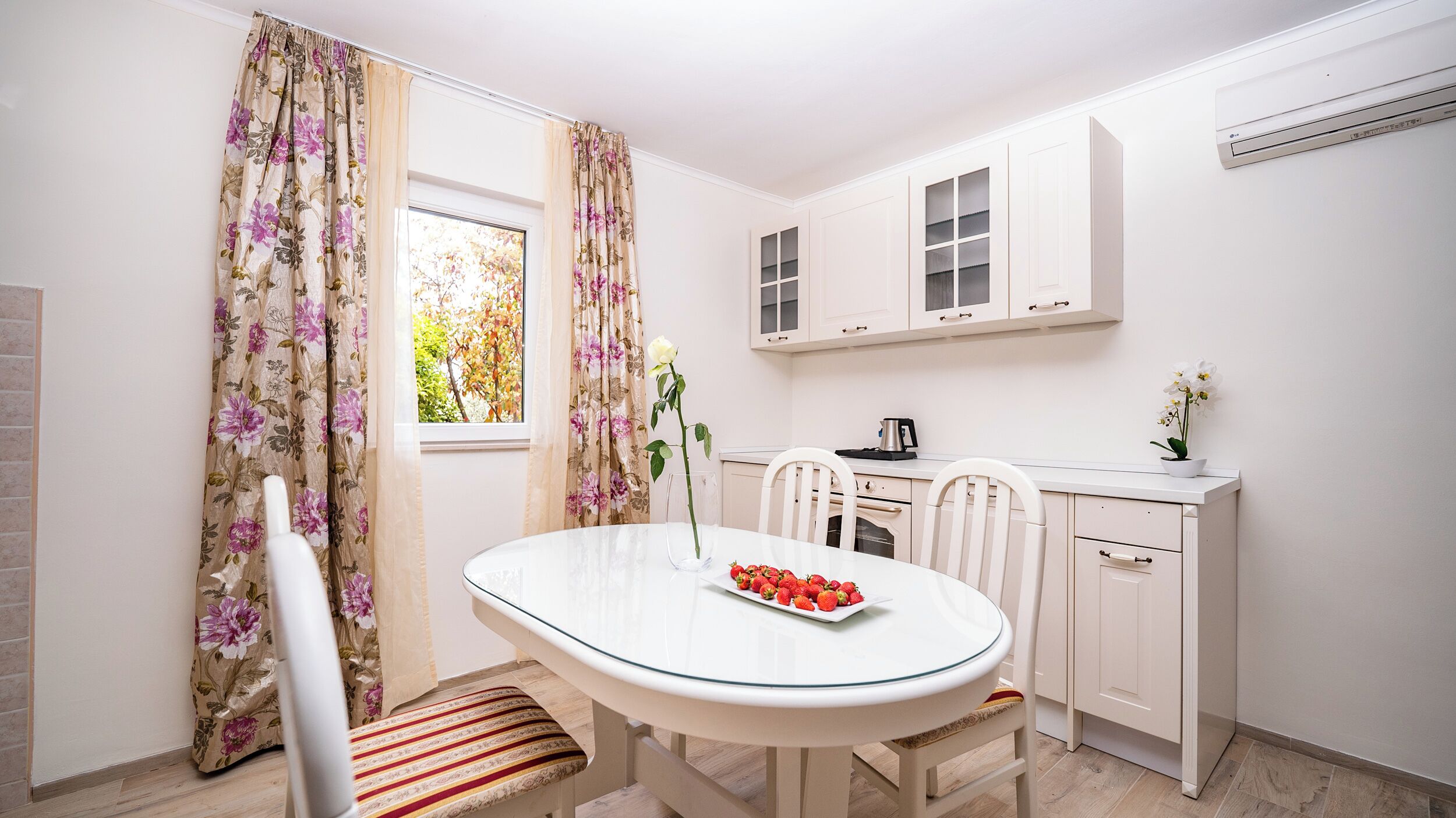 Cuina compacta amb armaris blancs, taula de dinar amb cadires i finestra amb cortines florals.