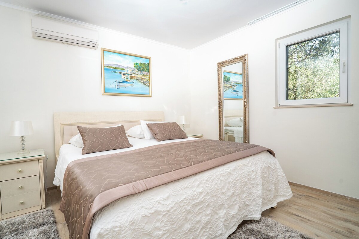 A bedroom with a bed, bedside table, lamp, air conditioning unit, paintings, and a window.