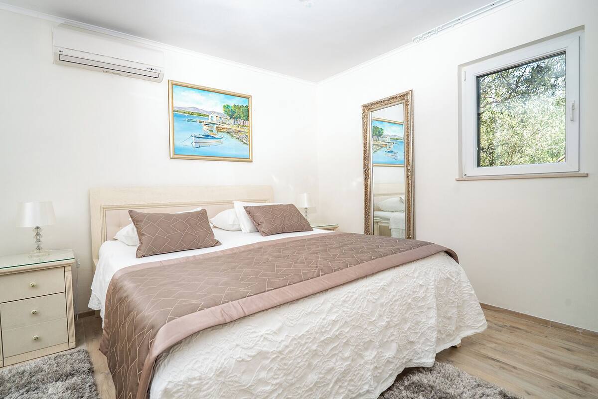 A bedroom with a bed, bedside table, lamp, air conditioning unit, paintings, and a window.