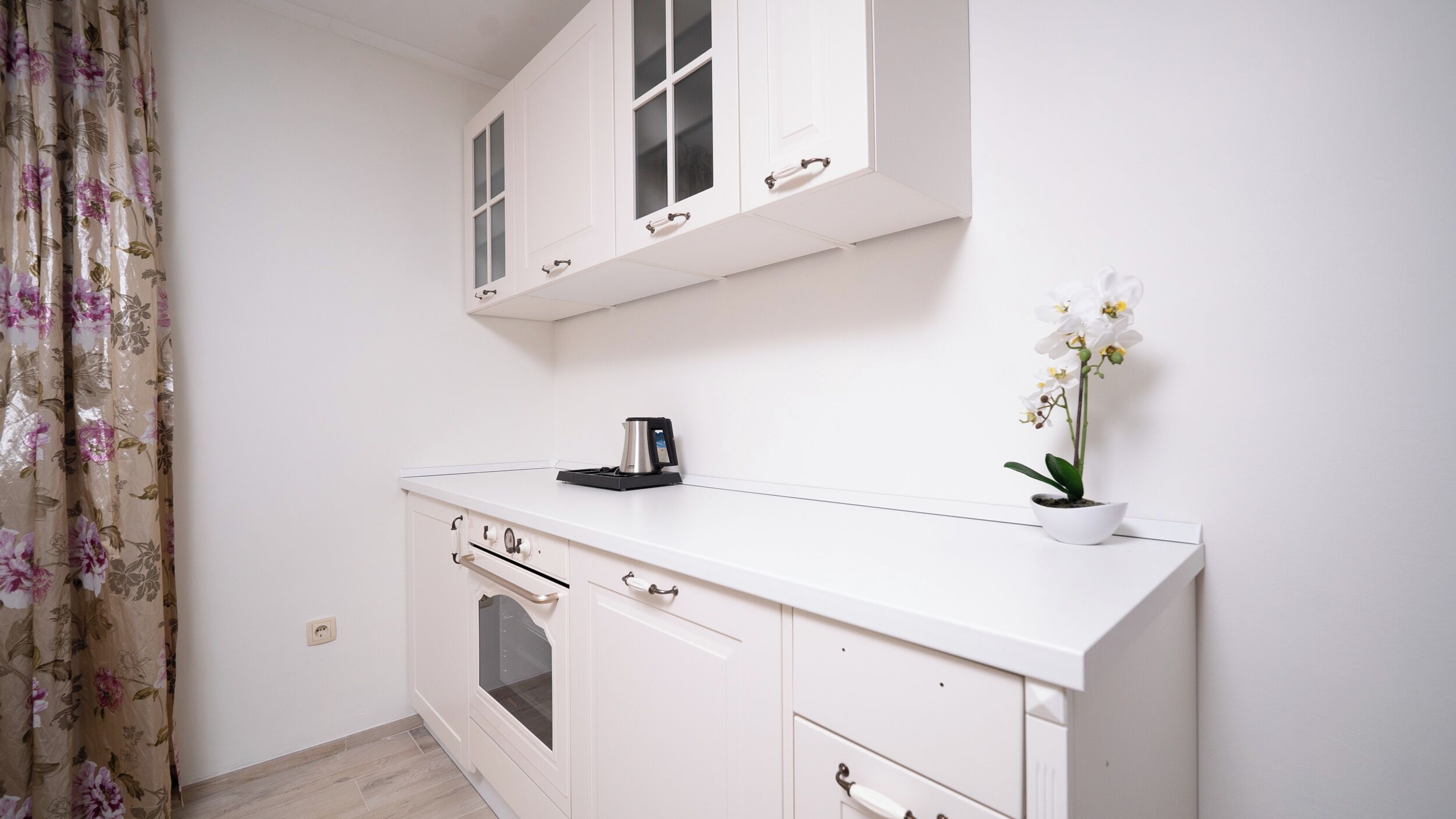 A white kitchen with a coffee maker, a potted orchid, and a window with blinds.