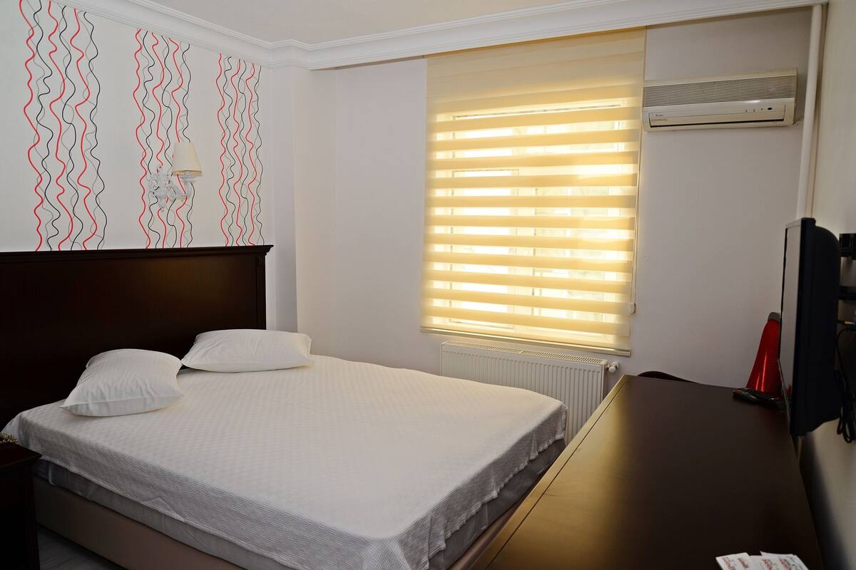 A hotel room with a bed, a desk, a radiator, and a window with blinds.