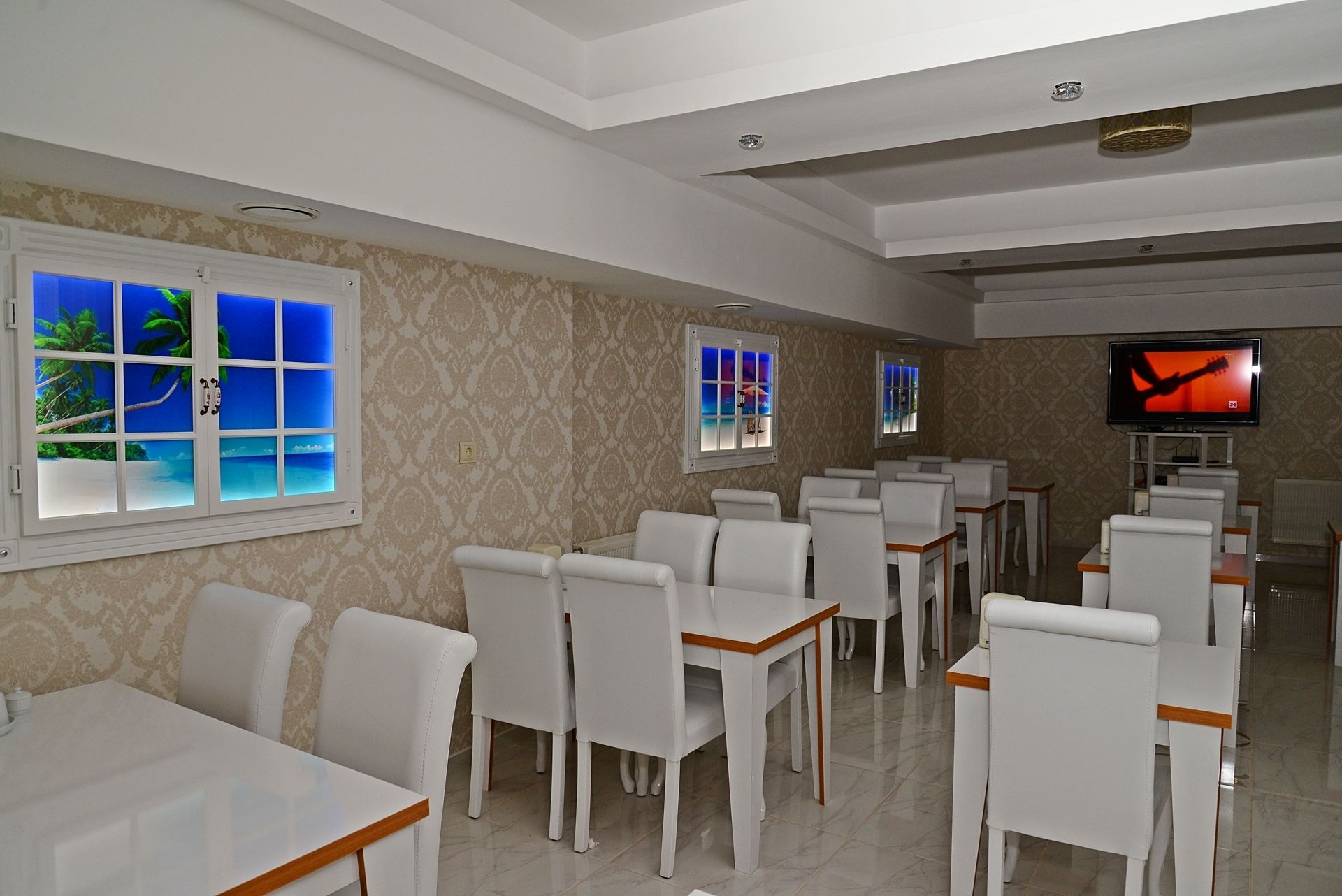 A restaurant with white chairs and tables, a TV on the wall, and windows with tropical scenes.