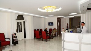 Un vestíbulo de hotel con una recepción, sillas rojas, un televisor y una escalera al fondo.