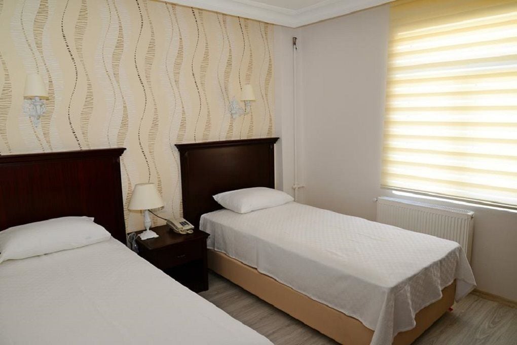 Una habitación de hotel con dos camas individuales, una mesita de noche con lámpara y una ventana con persianas.