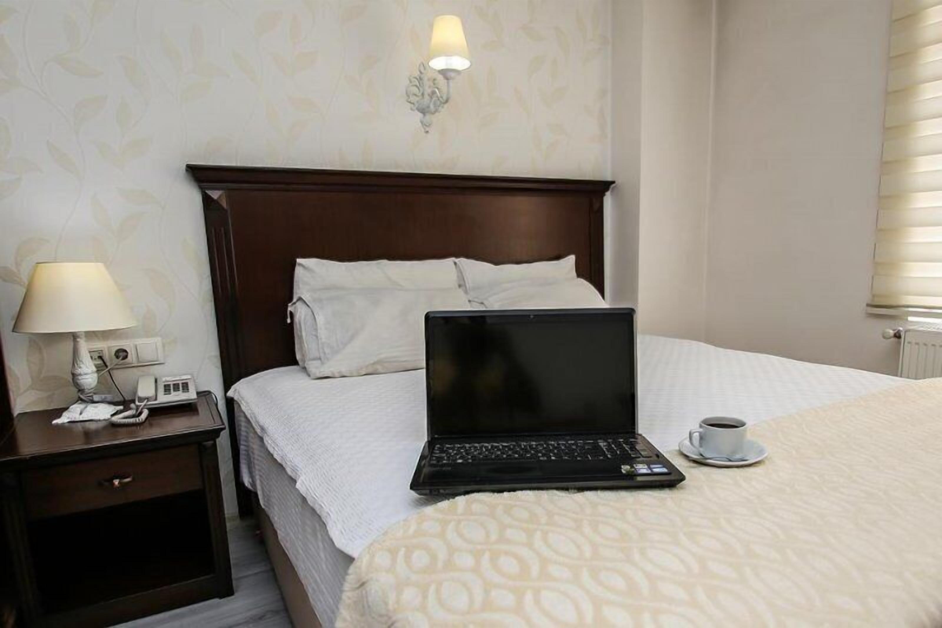 Una habitación de hotel con una cama, una mesita de noche con una lámpara, una computadora portátil y una taza de café sobre la cama.