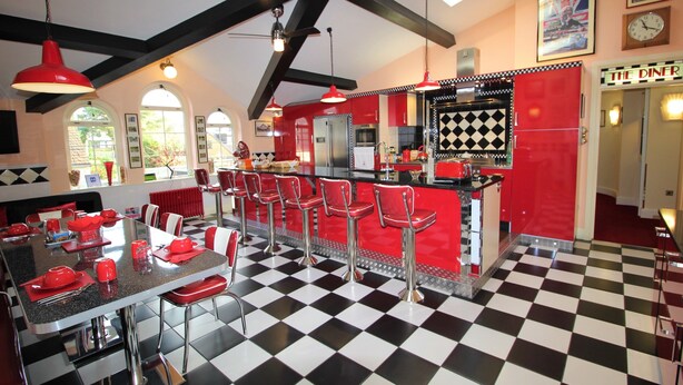A retro diner-style kitchen with checkered flooring, red bar stools, and a central island. - The PitStop (Bishop's Stortford)