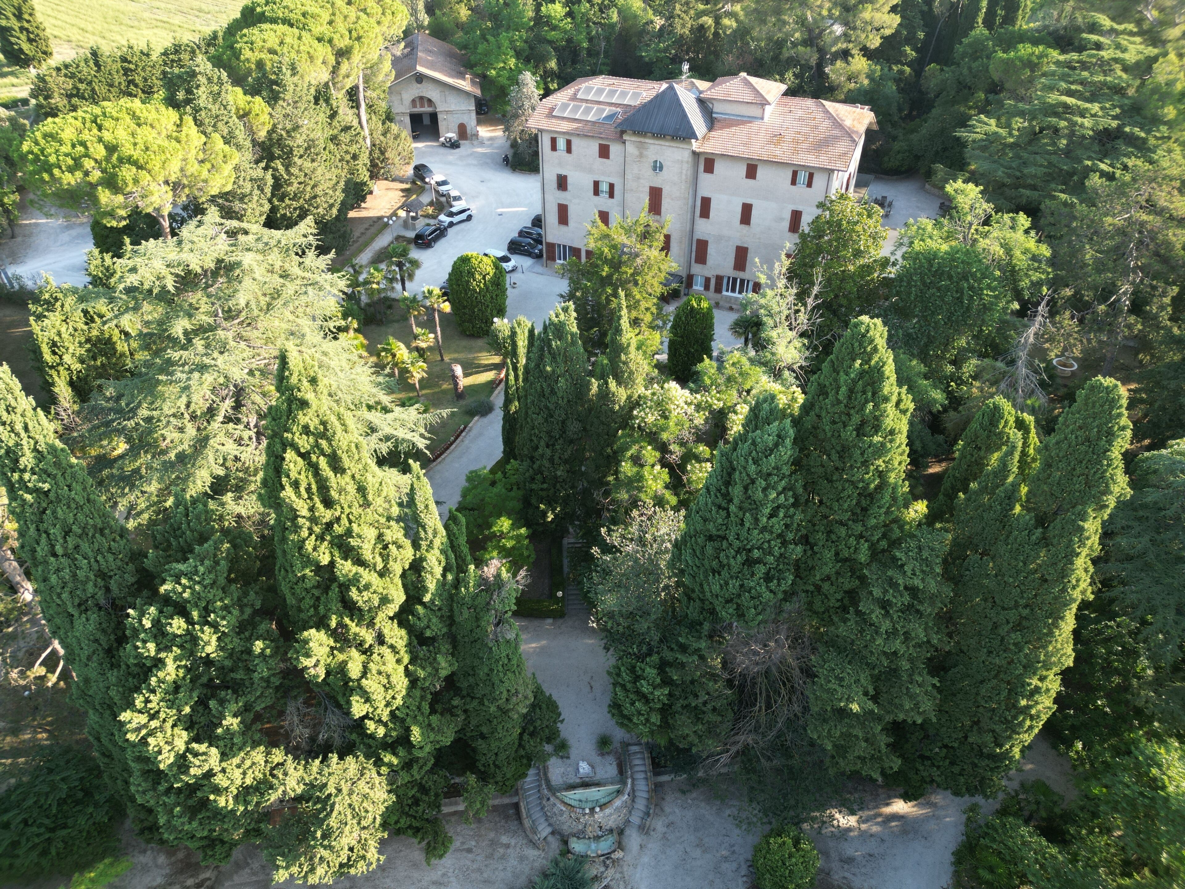 An aerial view of a large building surrounded by dense trees, with a parking area and a small garden.
