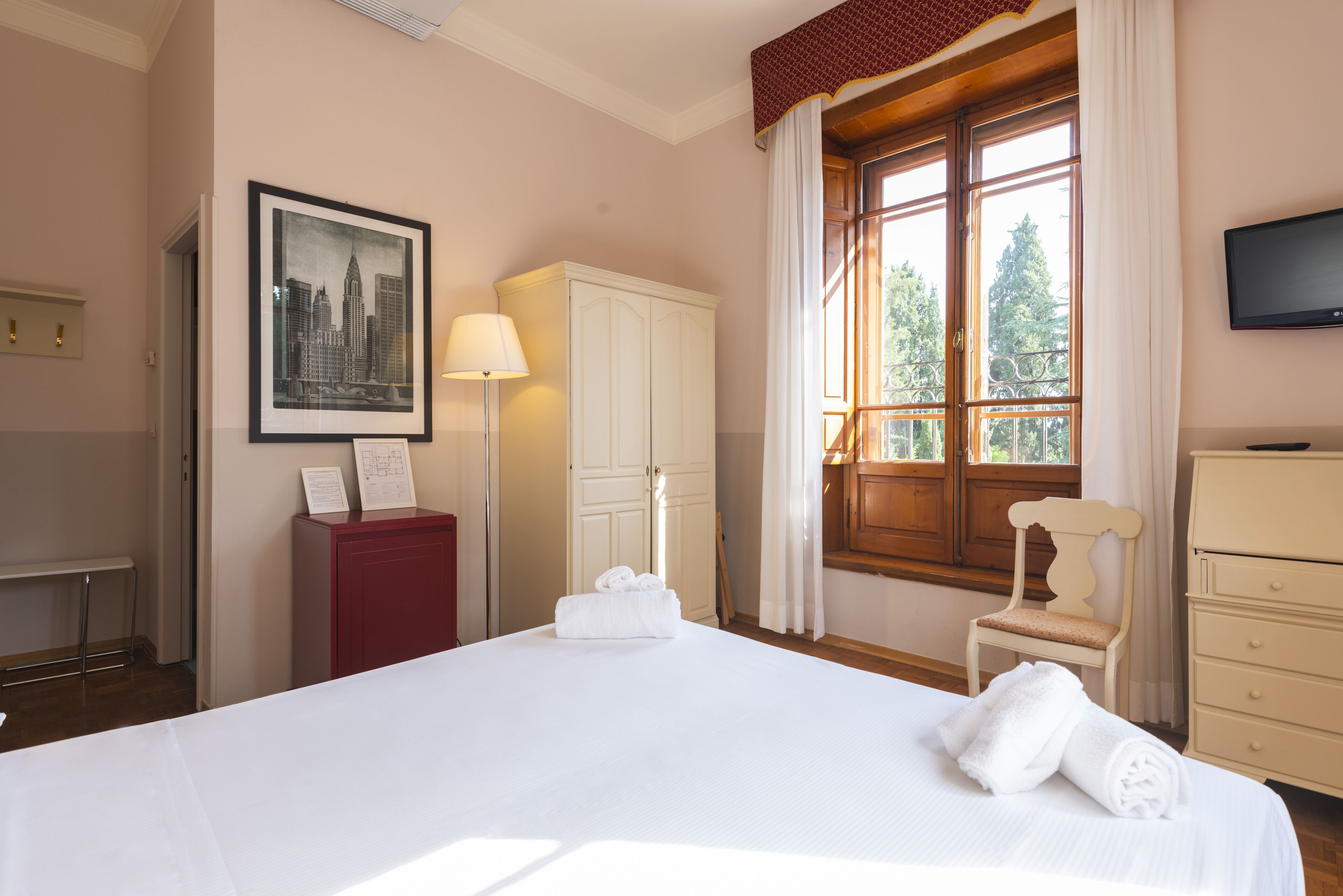 A hotel room with a large bed, a desk, a wardrobe, a chair, and a window with a view of trees.