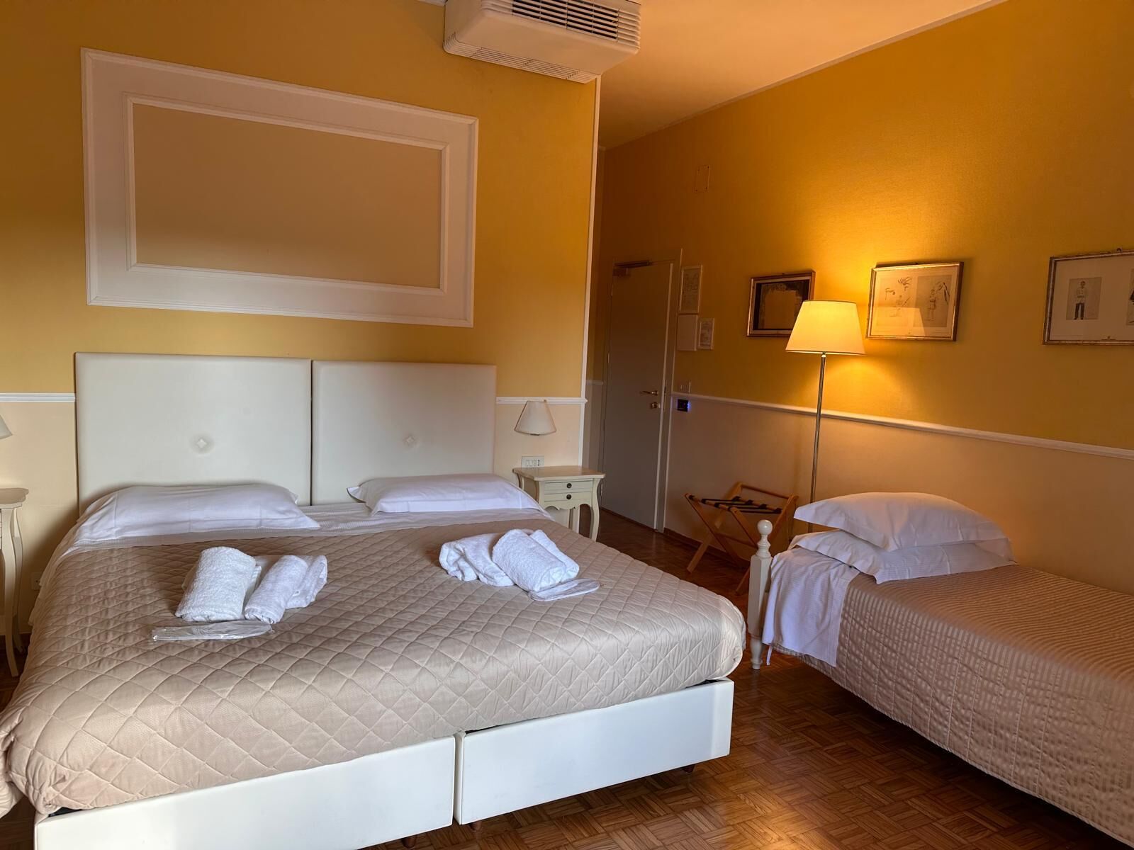 A hotel room with two beds, a bedside table, a lamp, and a wall-mounted air conditioner.
