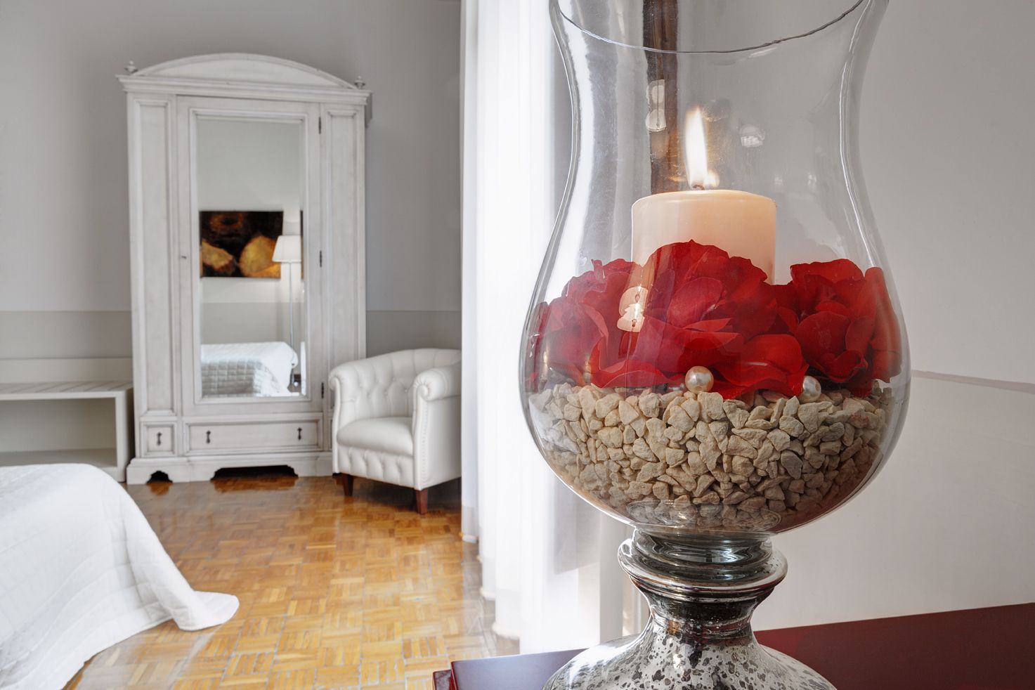 A room with a white armoire, a white armchair, and a glass table lamp with red flowers and stones.