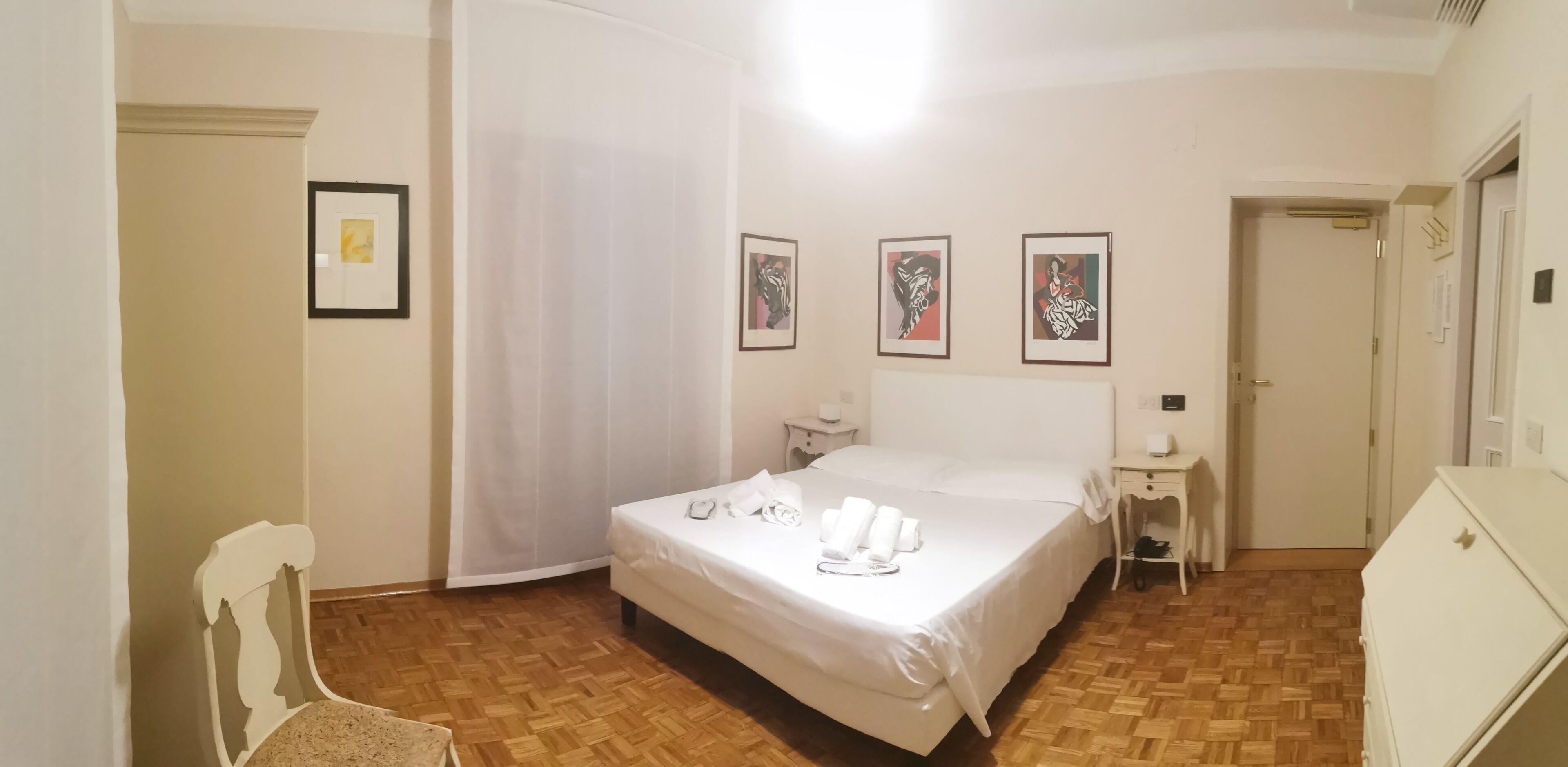Habitación de hotel con cama, mesita de noche y una silla. Se observan cuadros en la pared y una puerta a la derecha.