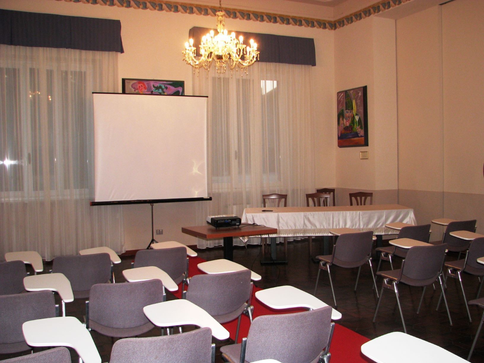 A conference room with a projector screen, rows of chairs, a table with a white tablecloth, and a chandelier.