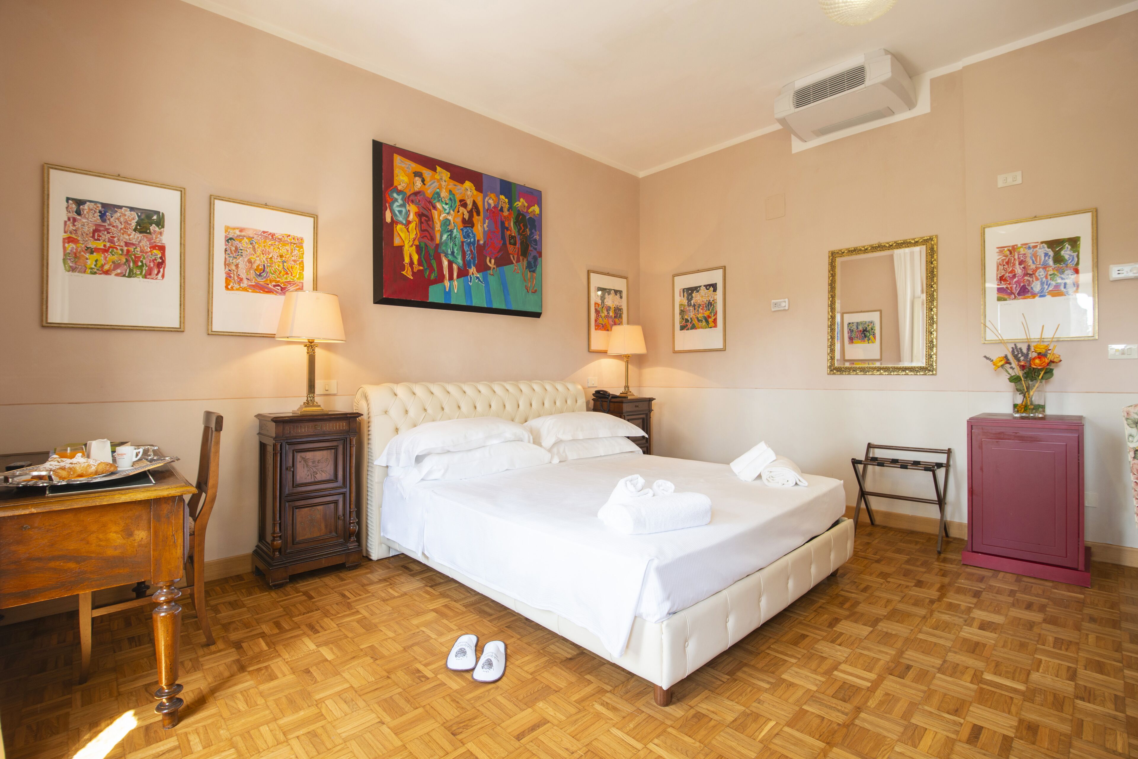 Habitación de hotel con una cama grande, dos mesitas de noche, un escritorio y una silla. Hay obras de arte enmarcadas en las paredes y un espejo sobre la cama.