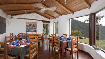 A dining area with wooden tables and chairs, a ceiling fan, and large windows offering a view of the outdoors.