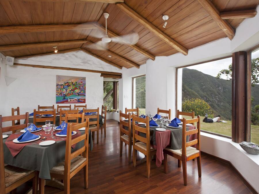 A dining area with wooden tables and chairs, a ceiling fan, and large windows offering a view of the outdoors.