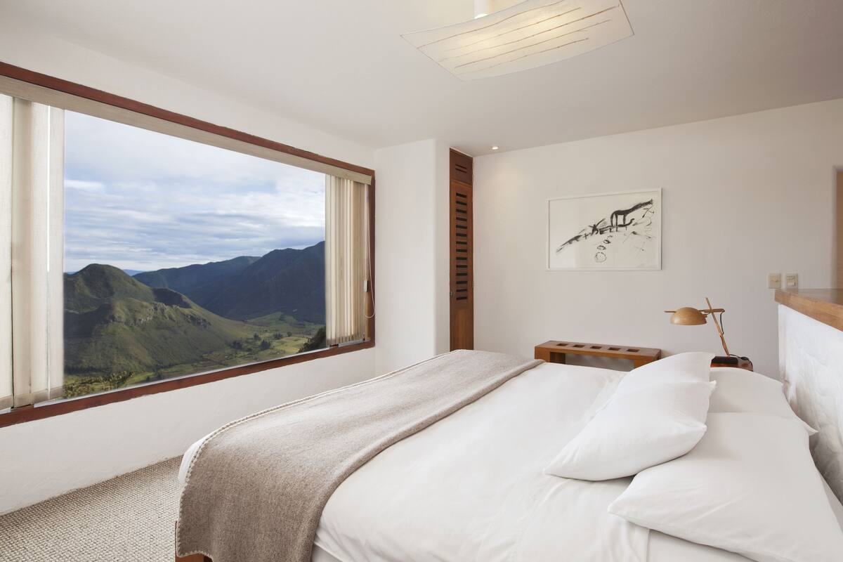A bedroom with a large window, a bed, a bedside table, a lamp, and a wall-mounted artwork.