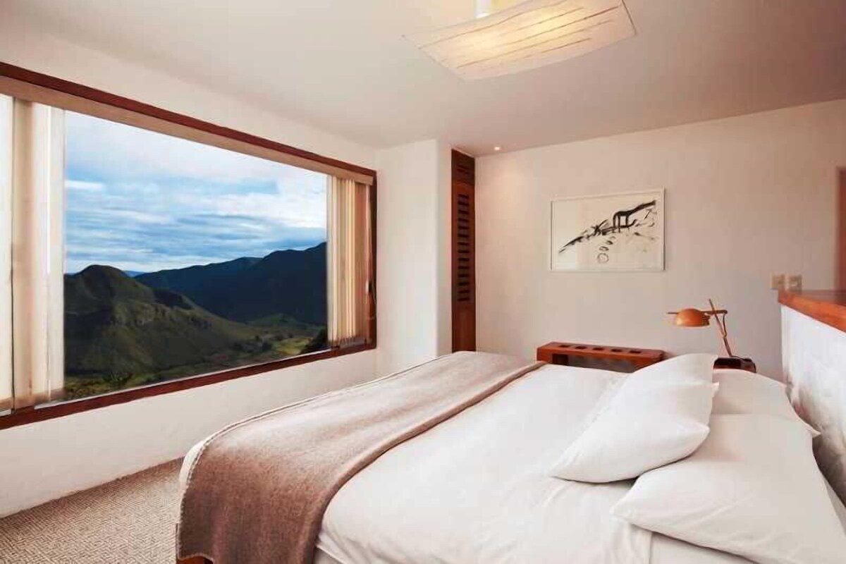 A bedroom with a large window offering a mountain view, a bed with white linens, a wooden headboard, and a wall-mounted artwork.