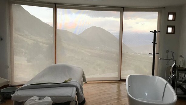 A room with a large sliding glass door leading to a mountainous landscape, a bathtub, and a massage table. - Hotel El Crater (San Antonio de Pichincha)