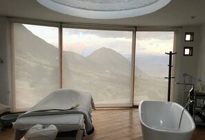 A room with a large sliding glass door leading to a mountainous landscape, a bathtub, and a massage table. - Hotel El Crater (San Antonio de Pichincha)
