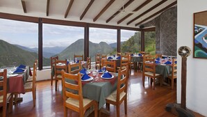 A restaurant with wooden tables and chairs, large windows offering mountain views, and a ceiling fan. - Hotel El Crater (San Antonio de Pichincha)