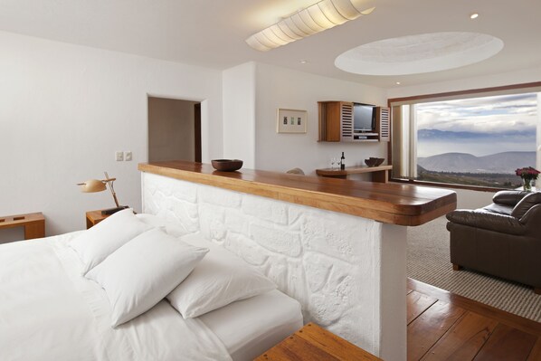A modern bedroom with a wooden headboard, a bed with white linens, a wooden nightstand, a desk, a television, and a view of mountains through a large window. - Hotel El Crater (San Antonio de Pichincha)