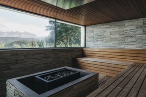 Um interior moderno com lareira de pedra, painéis de madeira e uma grande janela com vista para uma cadeia montanhosa coberta por floresta.