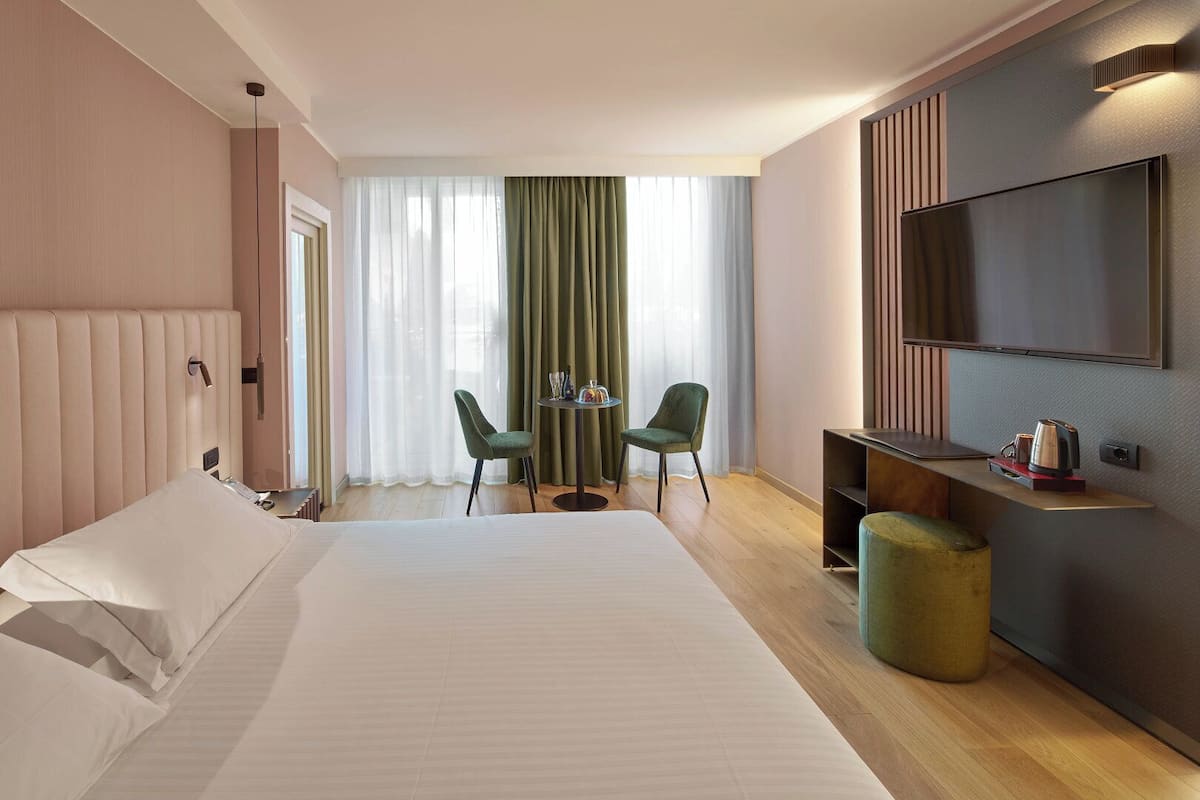 Una habitación de hotel con una cama grande, un escritorio con silla, televisor montado en la pared y una ventana con cortinas.