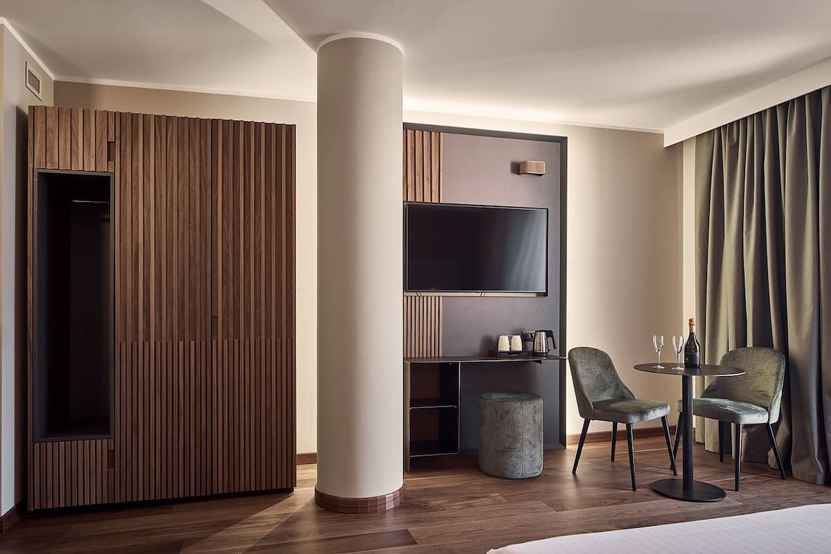 A modern hotel room with a wooden panel wall, a large flat-screen TV, a round coffee table, two chairs, and a large window with curtains.