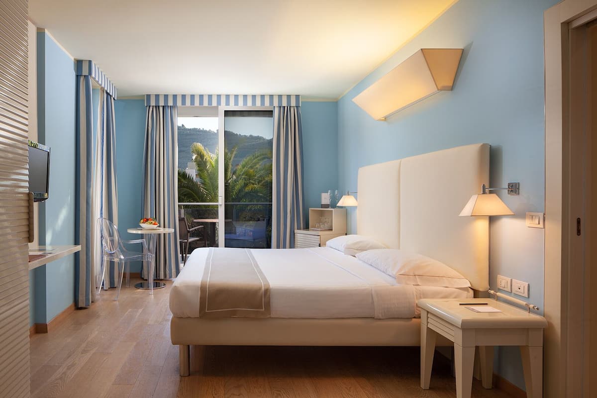 A hotel room with a bed, bedside tables, a TV, a balcony with curtains, and a view of the outdoors.