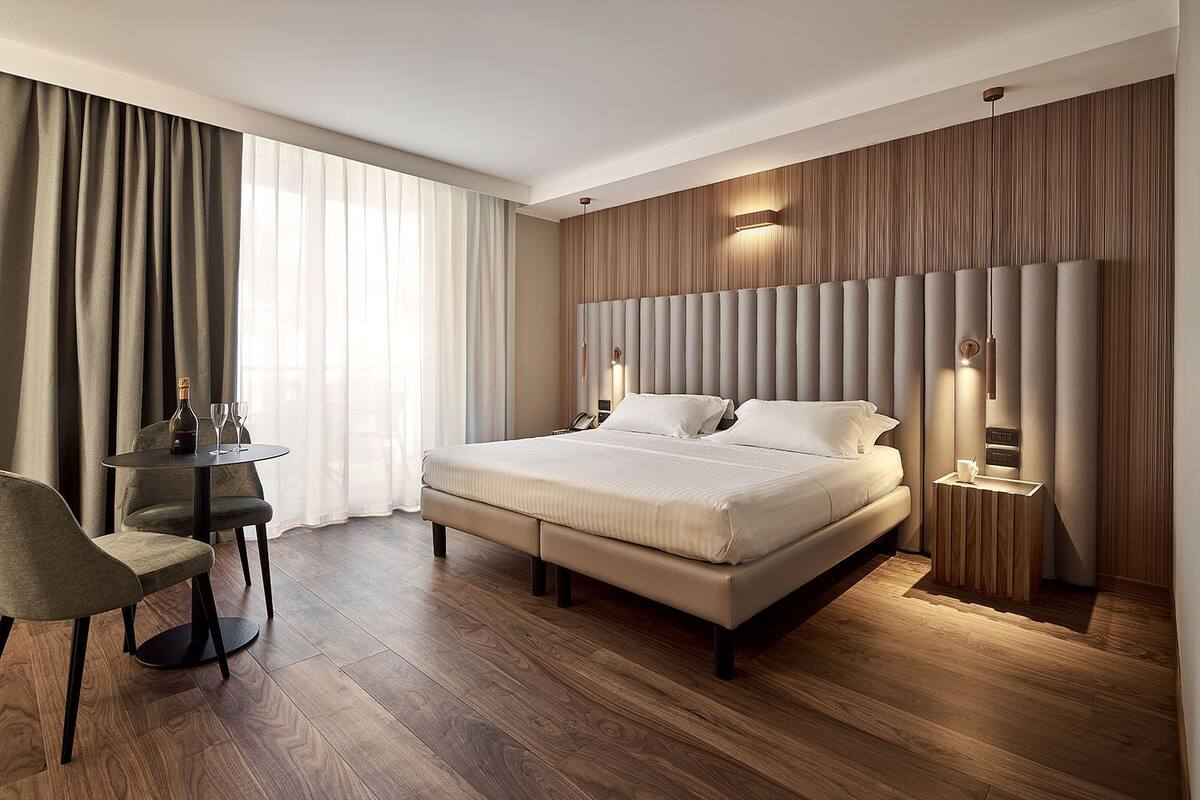 A modern hotel room with a large bed, a small table with a bottle and glasses, a chair, and a wooden floor.