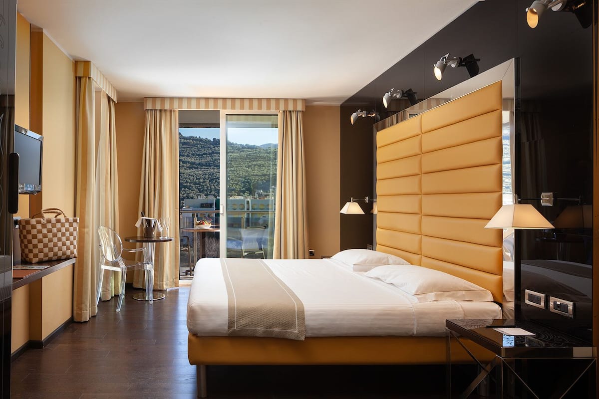 A hotel room with a large bed, a TV, a desk with a chair, a table with a glass of wine, and a view of a mountainous landscape through the window.