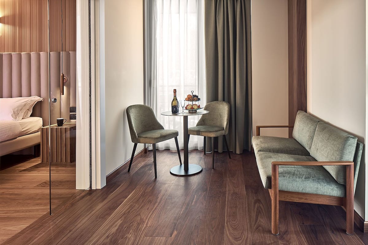 A modern hotel room with a sofa, two chairs, a small round table with a vase and fruit, and a bed.