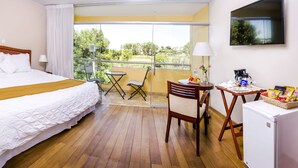 A hotel room with a large bed, a small table with a chair, a refrigerator, and a balcony with a view of greenery. - Alwa Premium Hotel Boutique (Arequipa)