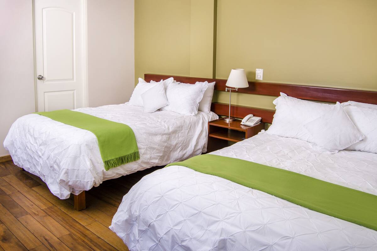 Twee eenpersoonsbedden met witte lakens en groene plaids, een houten nachtkastje met een lamp en een witte deur.