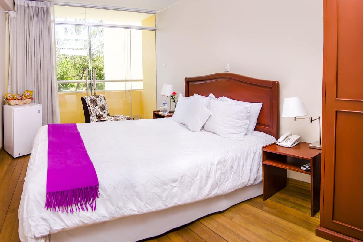 Hotelkamer met een groot bed, een houten hoofdeinde, een nachtkastje met een lamp, een tafeltje met snacks, een stoel en een raam met gordijnen.