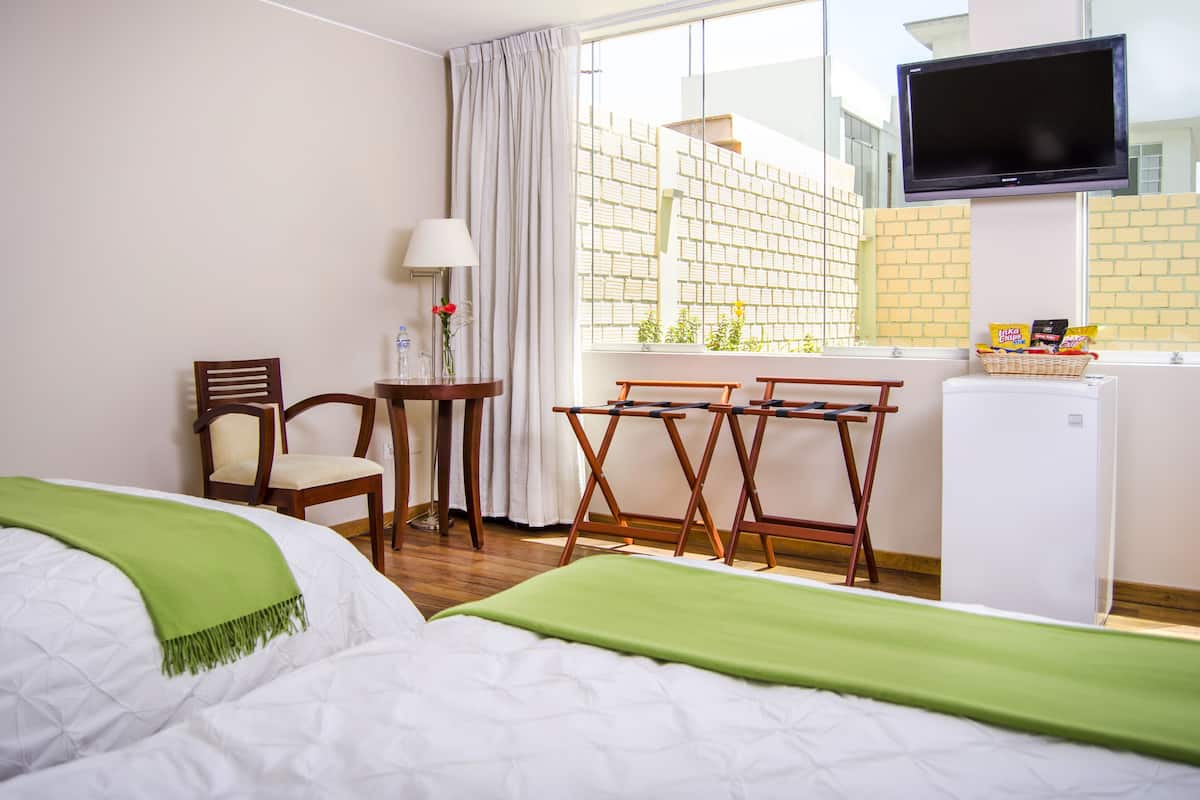 Een hotelkamer met een bed, een stoel, een klein tafeltje met een lamp, een aan de muur bevestigde televisie en een raam met uitzicht op gebouwen.