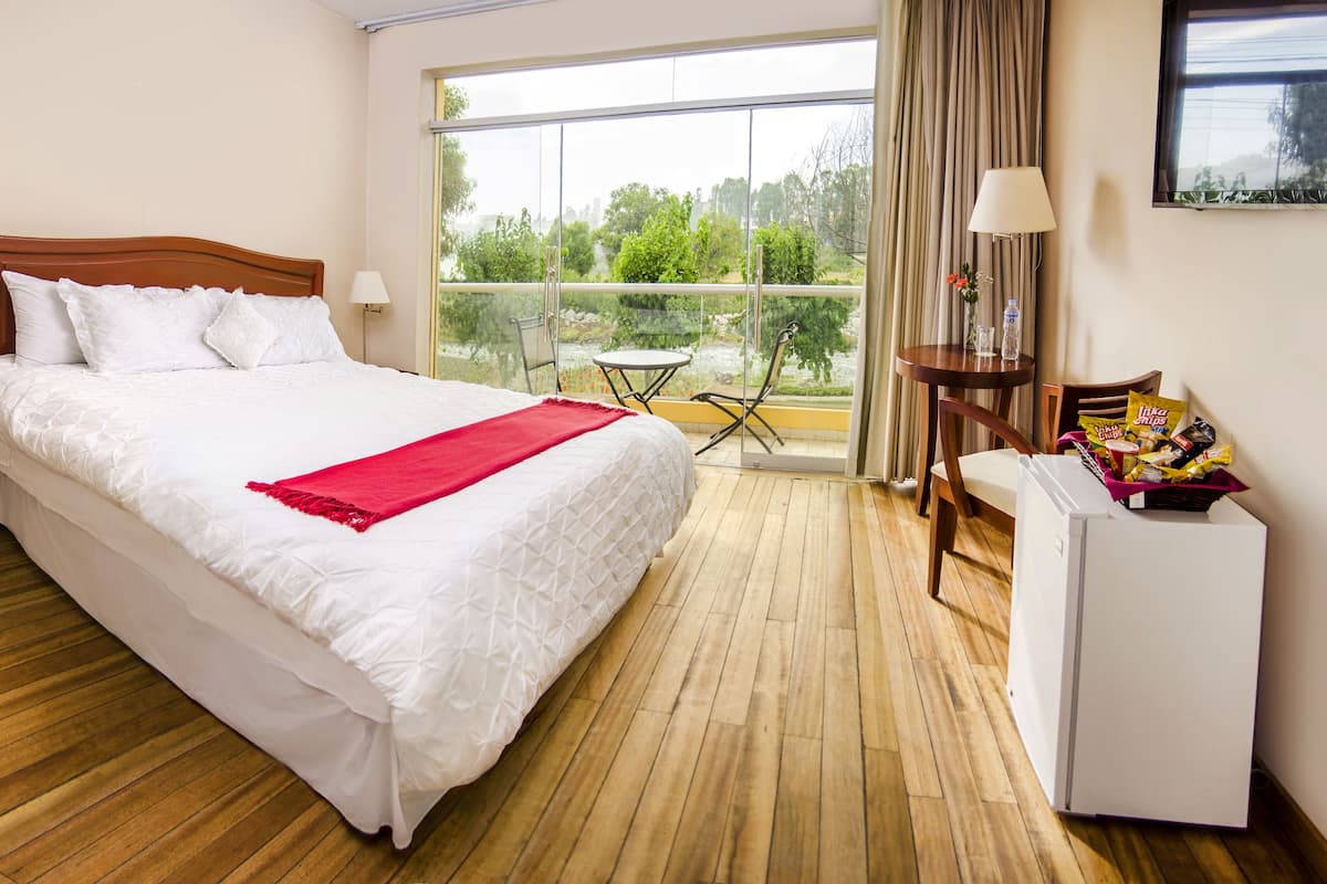 Een hotelkamer met een groot bed, een bureau met stoel, een kleine koelkast, en een balkon met uitzicht op groen.