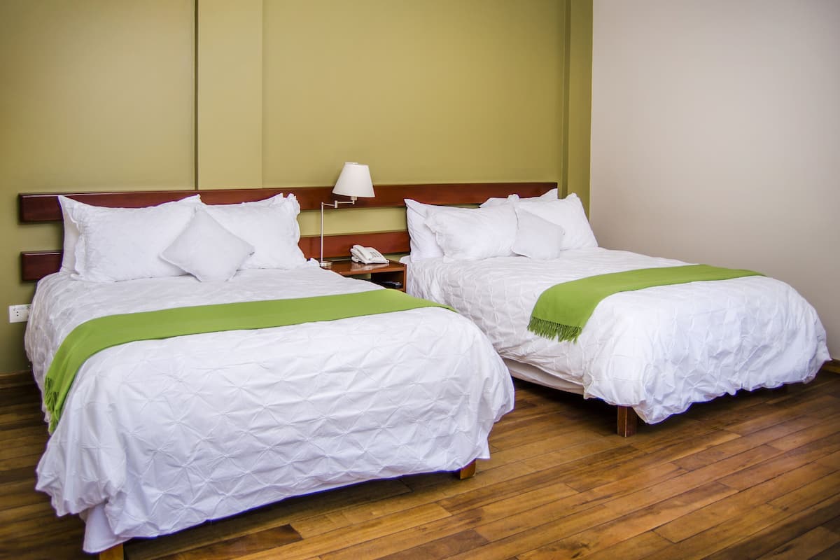Twee eenpersoonsbedden met witte lakens en groene plaids, houten hoofdeinden en een nachtkastje met een lamp en telefoon.