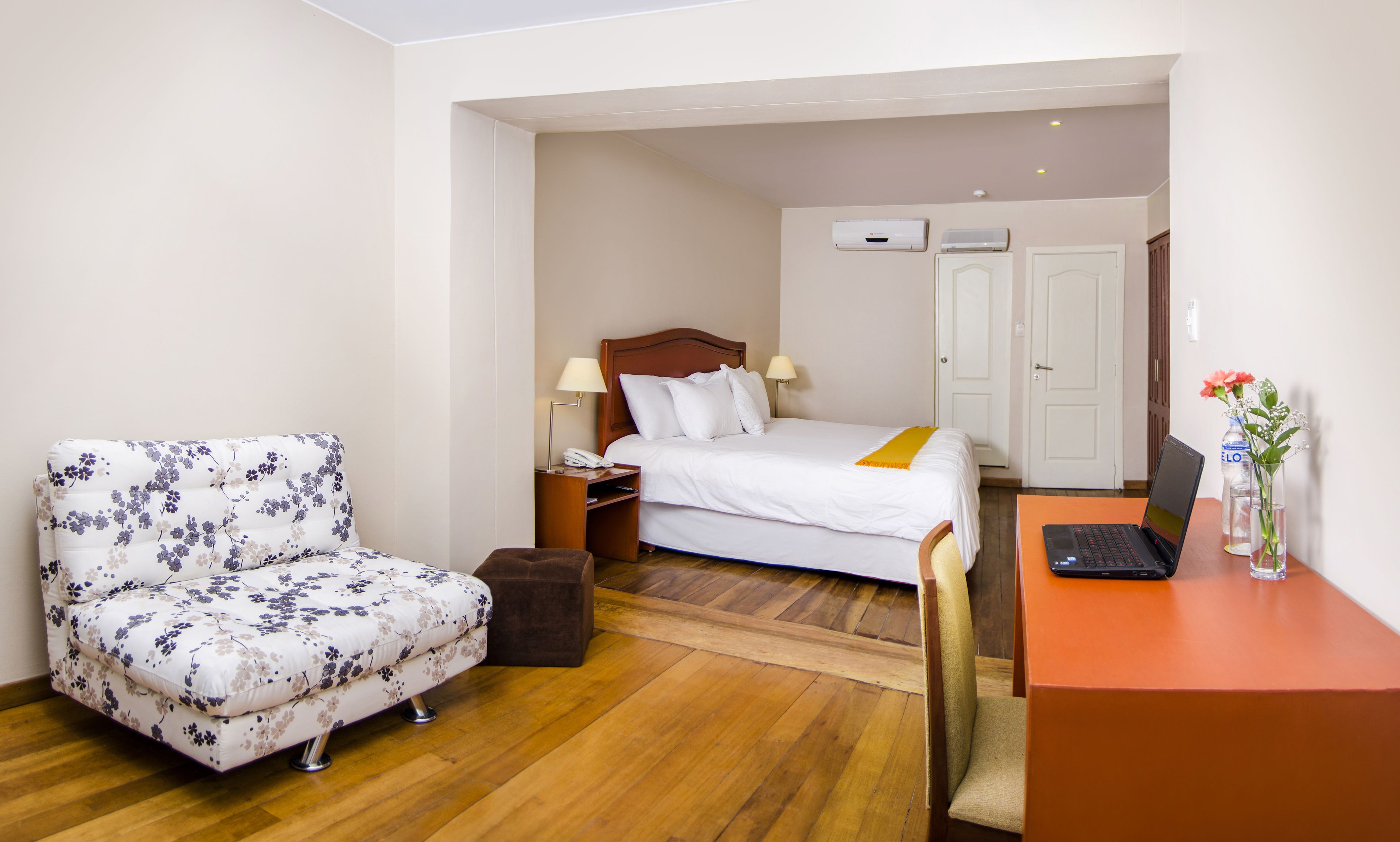 Habitación de hotel con cama, escritorio con computadora portátil, silla, mesita de noche con teléfono y una pequeña mesa con un jarrón de flores.