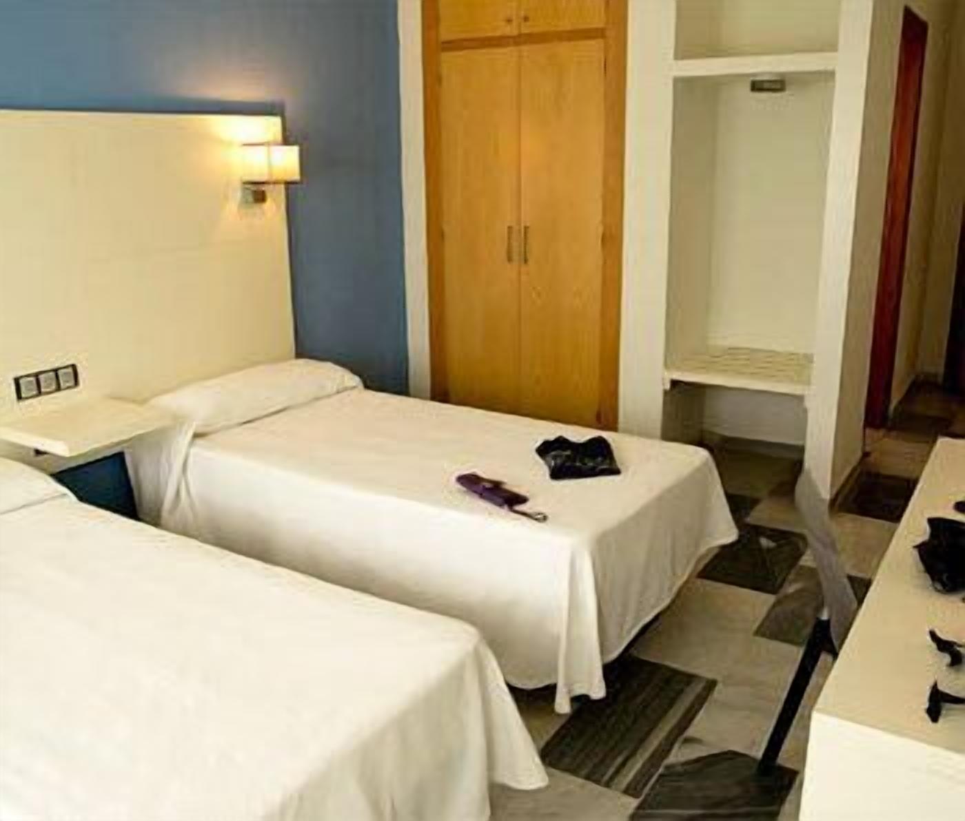 A hotel room with two beds, a wooden wardrobe, a small desk, and a shelf.