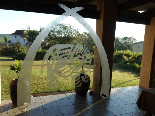 A frosted glass cutout of a fish with a cross, potted plants, and a view of a green lawn and houses in the background. - Elephant's Nest Guest House (Graskop)