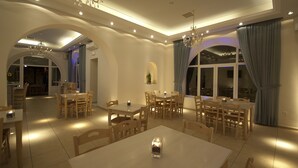 Un amplio comedor con varias mesas y sillas, un candelabro y ventanas grandes con cortinas.