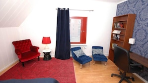 A room with a red sofa, two blue armchairs, a red lamp, a black office chair, a bookshelf, and a window with a red frame. - Avesta Stadshotell (Avesta)
